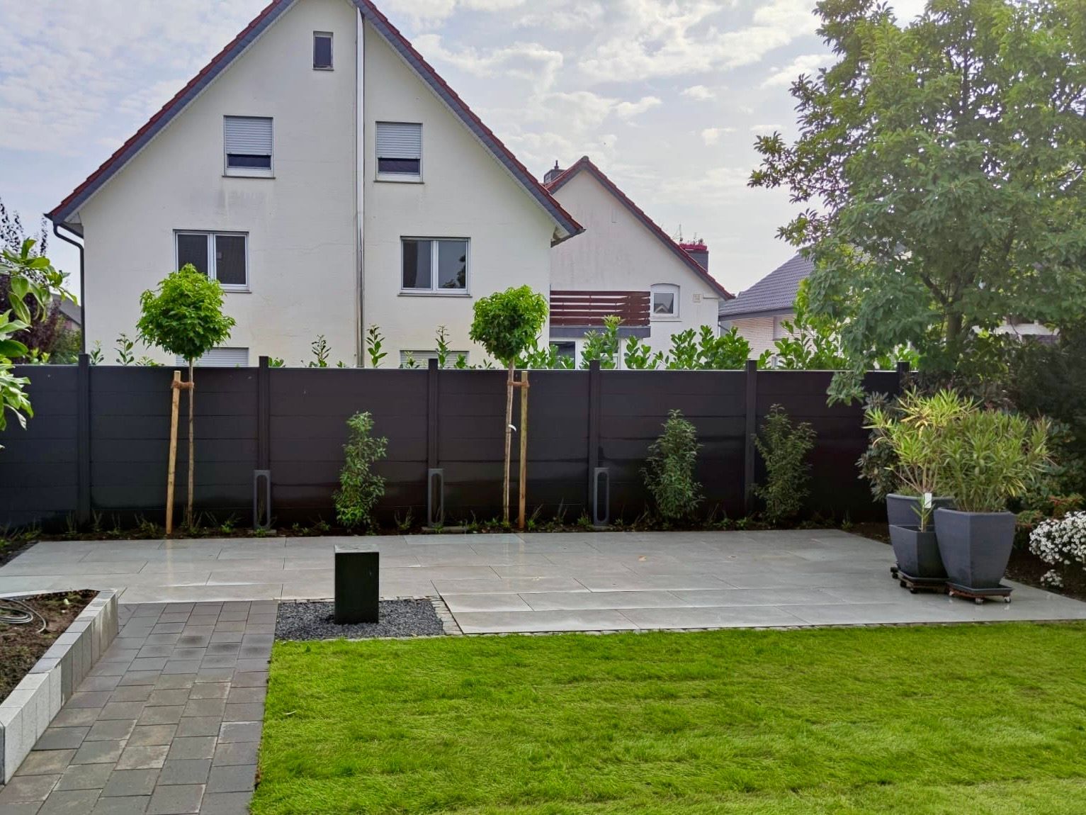 Hinterhof mit grauer Terrasse, Rasen, schwarzem Zaun, Bäumen und zweistöckigen Häusern im Hintergrund.