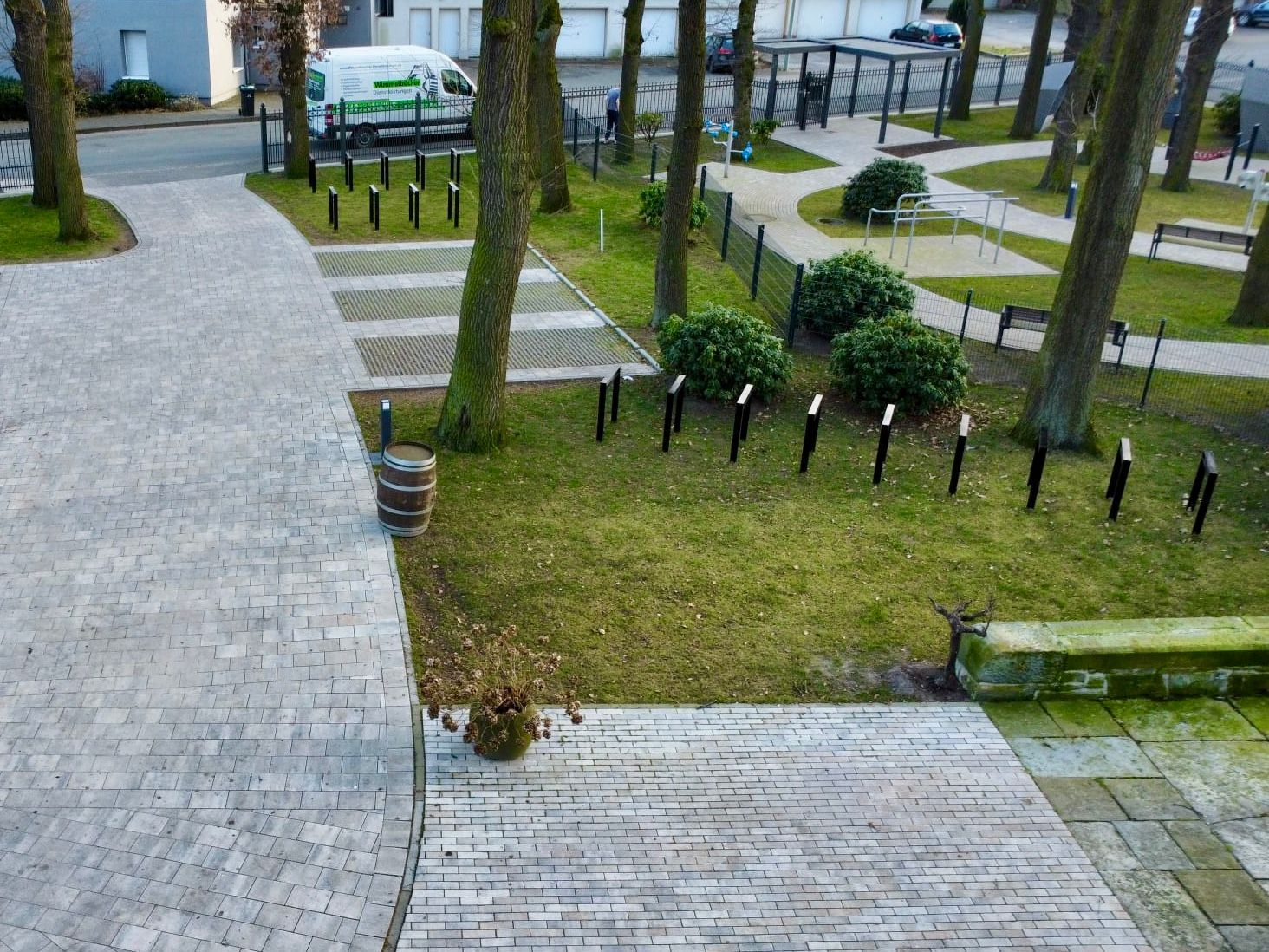Blick auf einen Park mit Bäumen, Gras, Bänken und gepflasterten Gehwegen. Auf einer nahegelegenen Straße ist ein Fahrzeug geparkt.