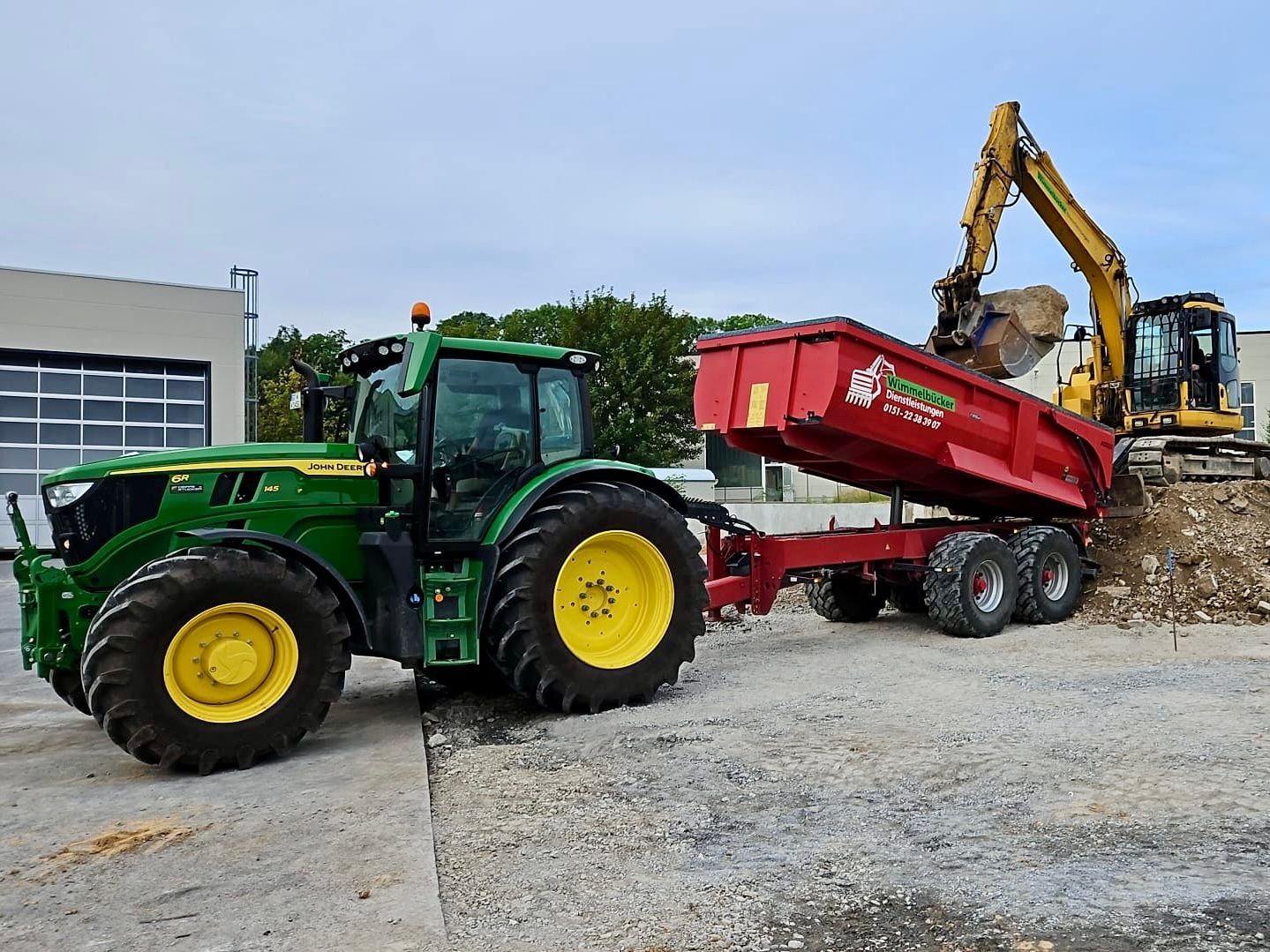 Ein John Deere-Traktor zieht einen roten Anhänger, während ein Bagger den Anhänger mit Kies belädt.