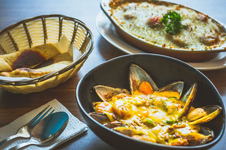 Tres platos sobre la mesa: un guiso al horno, mejillones en salsa y pan en una cesta de mimbre.