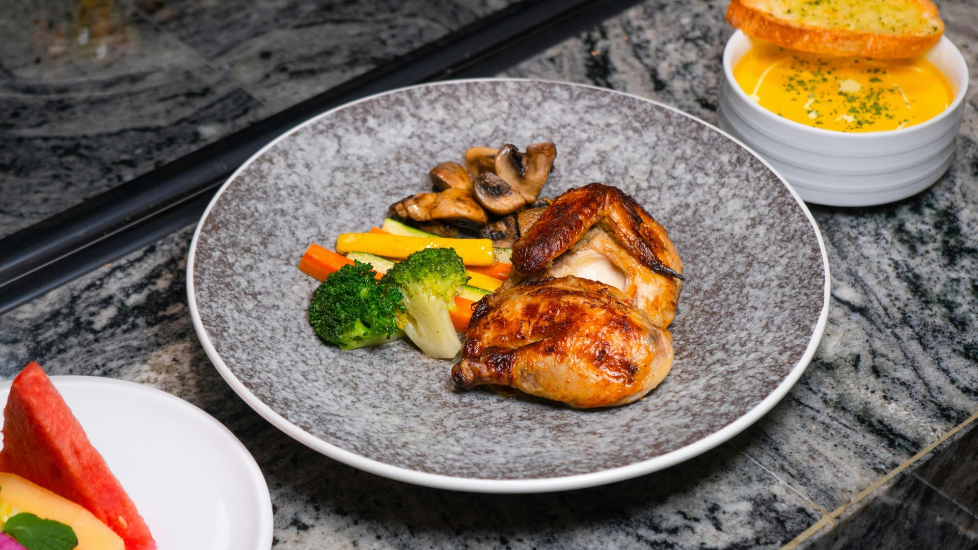 Pollo a la parrilla con brócoli y champiñones en un plato gris junto a un cuenco blanco con salsa.
