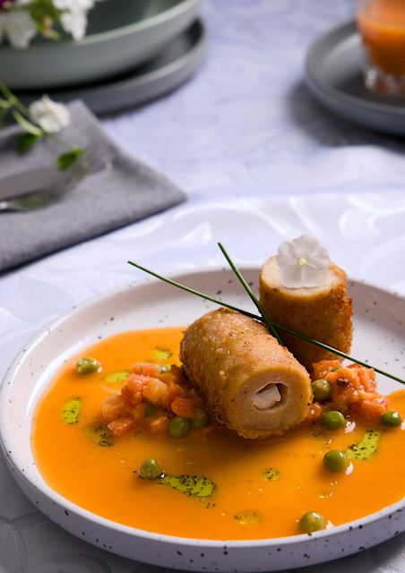 Dos croquetas empanizadas en salsa de naranja, adornadas con cebollino y crema, servidas en un plato blanco.
