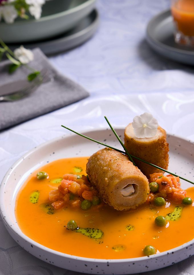 Dos croquetas empanizadas en salsa de naranja, adornadas con cebollino y crema, servidas en un plato blanco.