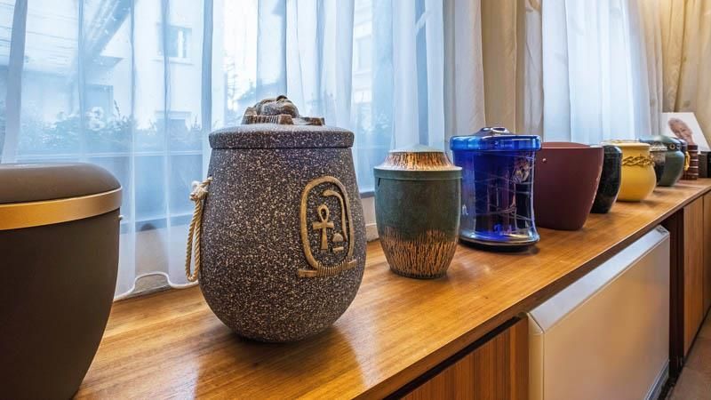 Eine Reihe von Urnen steht auf einem Holzregal vor einem Fenster.