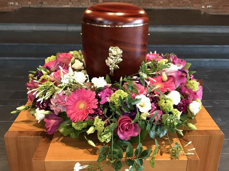 Eine Holzurne ist von rosa und weißen Blumen umgeben und steht auf einem Holztisch.