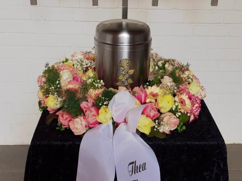 Eine Urne ist von Blumen und einem Band mit der Aufschrift „theu“ umgeben