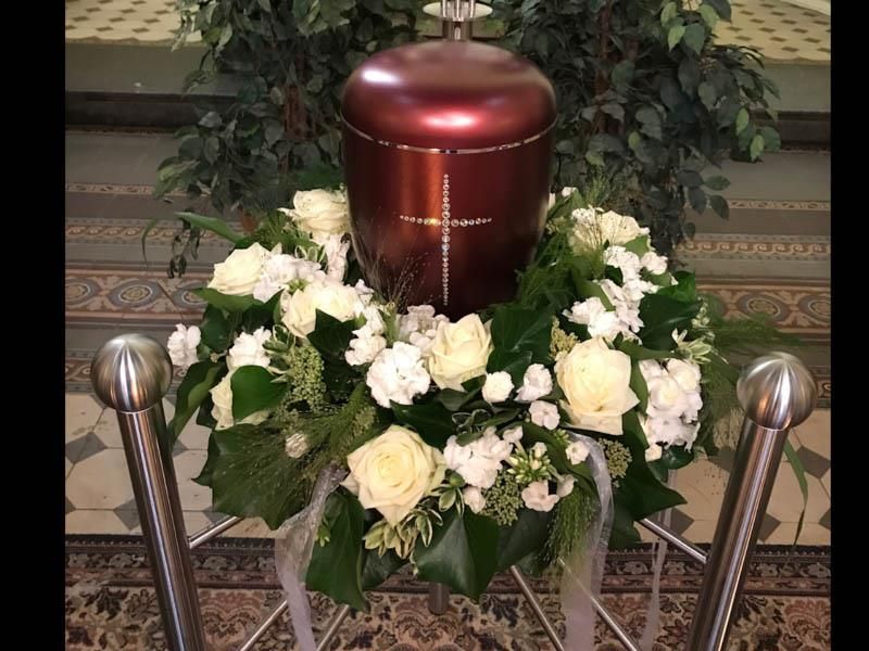 Eine rote Urne ist von weißen Rosen und Grün umgeben.