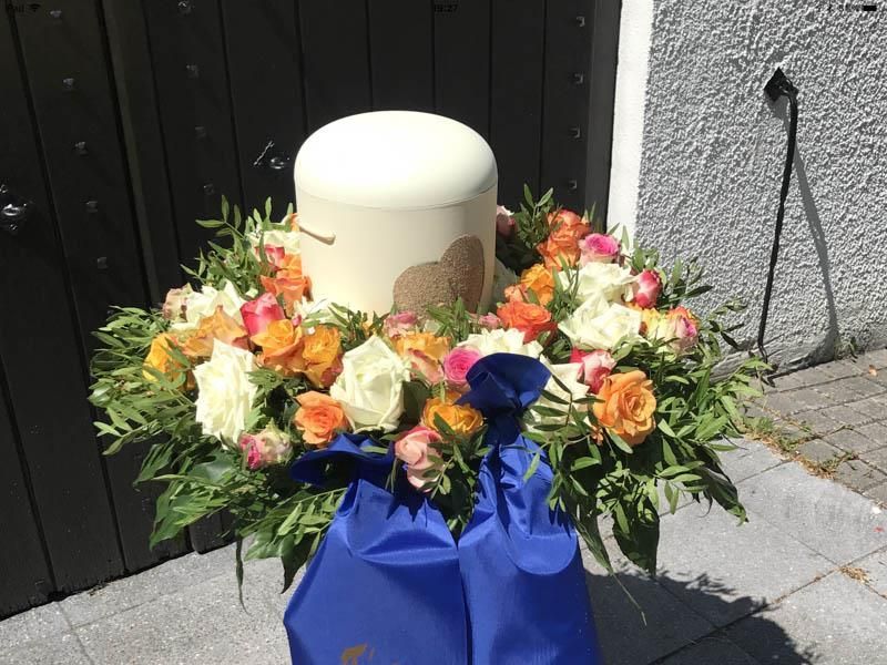 Eine weiße Urne ist von Blumen und einer blauen Tasche umgeben