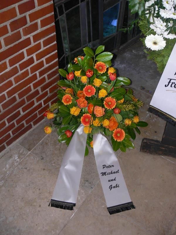 Ein Kranz aus orangen und gelben Blumen mit einem weißen Band, auf dem 