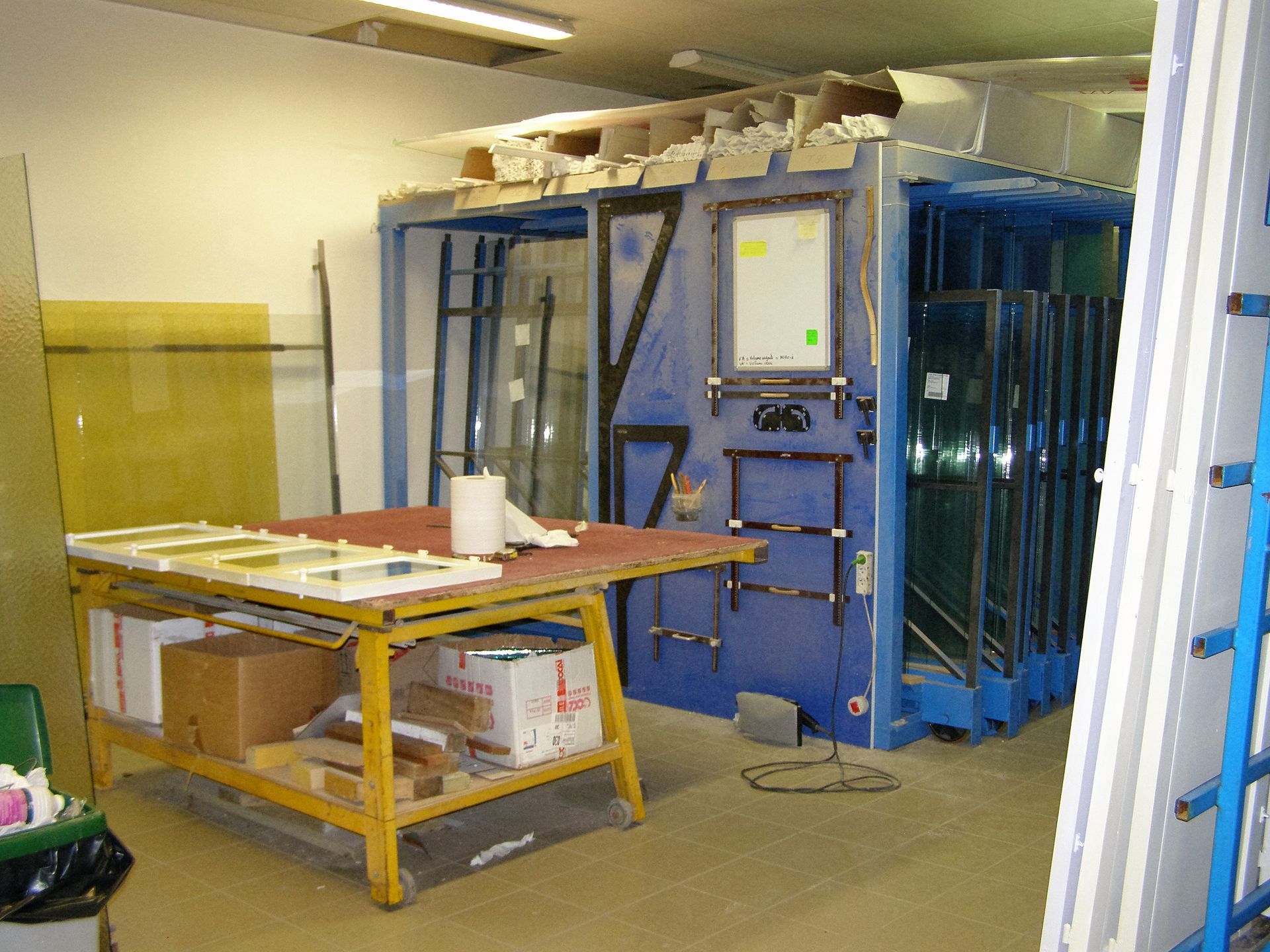 Un établi jaune se trouve à côté d'un rayonnage industriel bleu supportant de grandes plaques de verre.