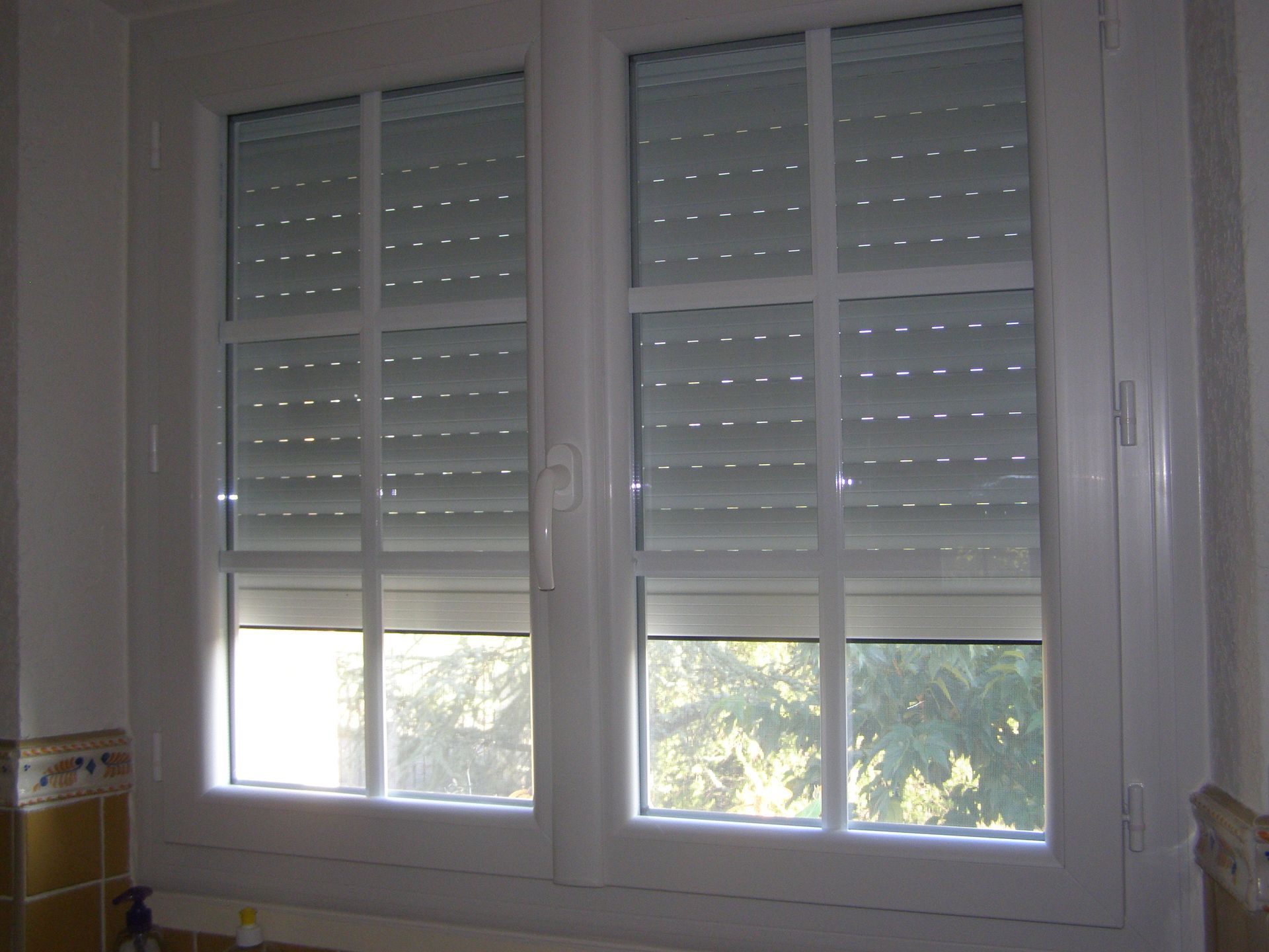 Une fenêtre à cadre blanc et six carreaux, partiellement recouverte de volets roulants ajourés.