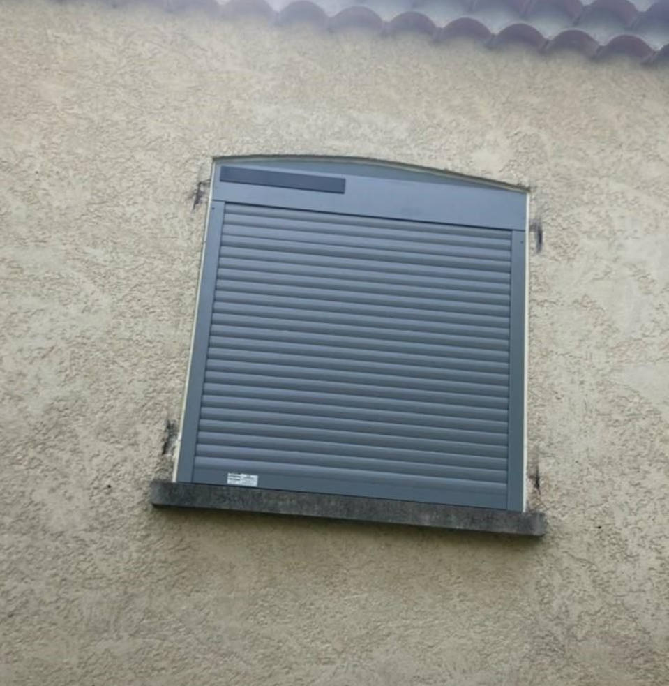 Un volet roulant gris, entièrement fermé, sur un mur extérieur en stuc clair.