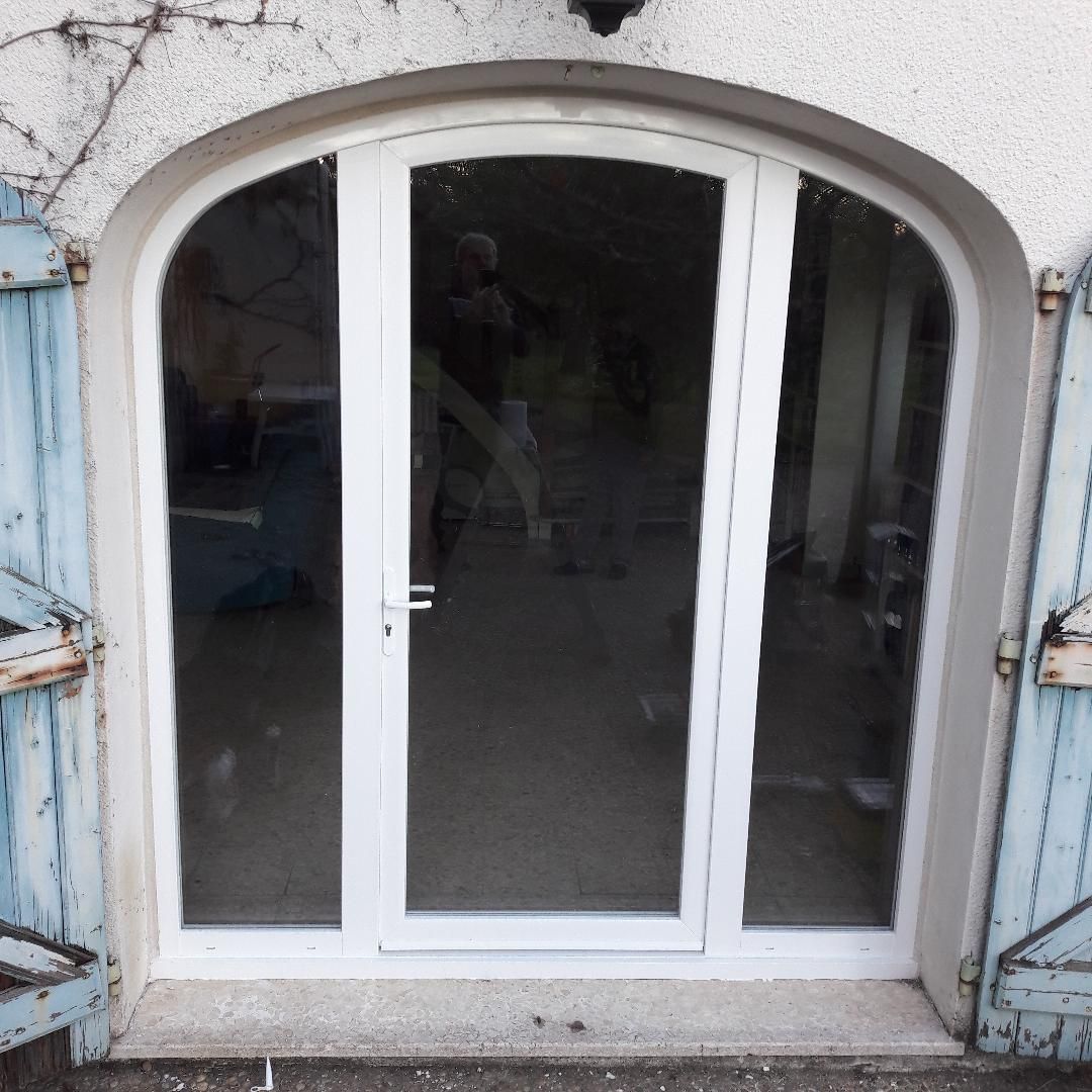 Une porte vitrée blanche cintrée, encadrée par une porte en pierre et flanquée de volets bleus.