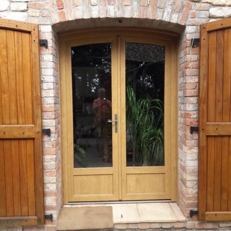 Deux portes vitrées à cadre en bois clair, placées dans une arche en briques et flanquées de volets en bois.