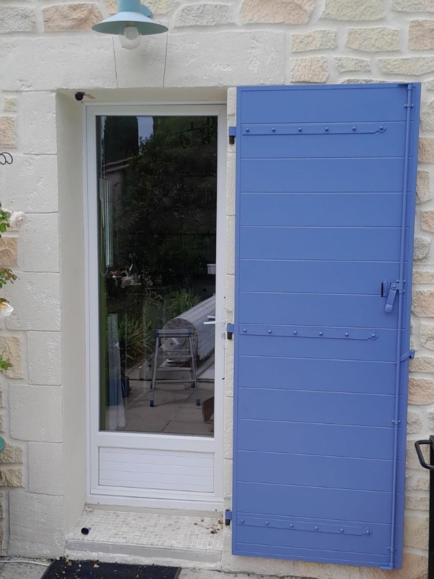 Une porte vitrée blanche encastrée dans un mur de pierre, avec un volet en bois bleu.