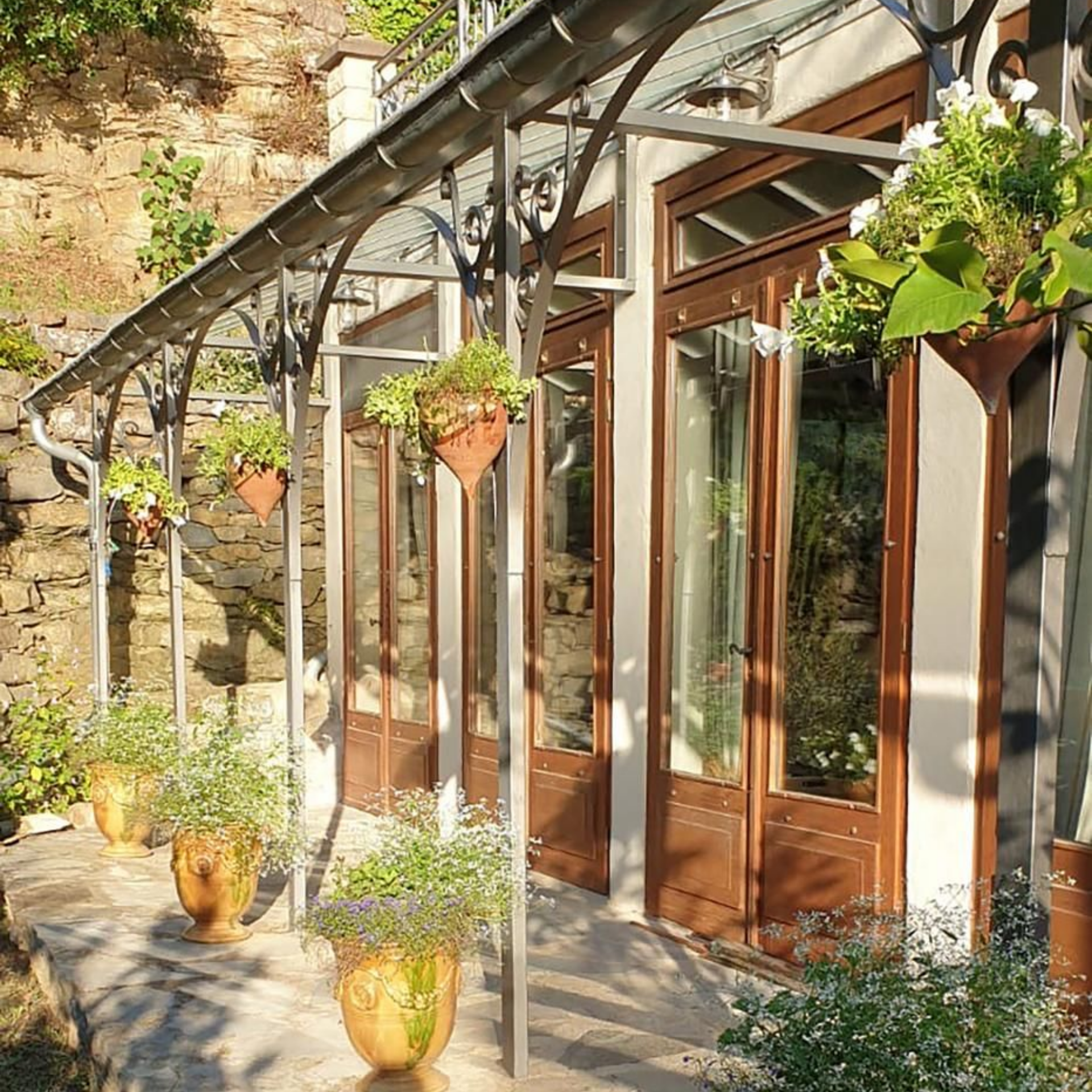 Une terrasse en pierre, sous une treille en métal fixée à un bâtiment aux hautes portes vitrées.