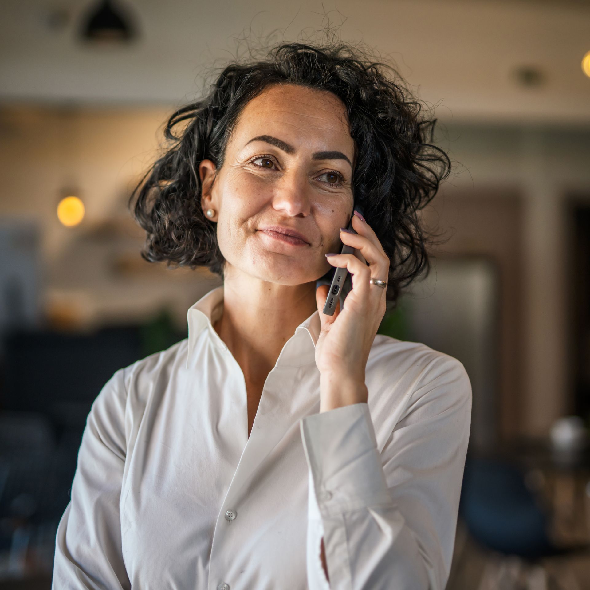 Une personne aux cheveux bouclés et foncés, vêtue d'une chemise blanche à col, tient un smartphone à l'oreille.