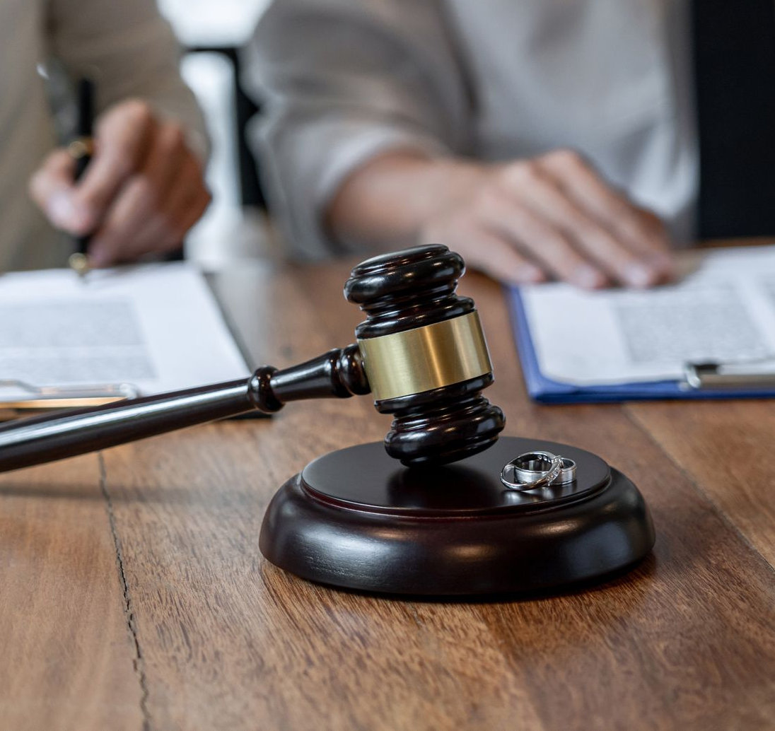 Un maillet en bois et deux alliances posés sur un bureau, avec des personnes examinant des documents juridiques