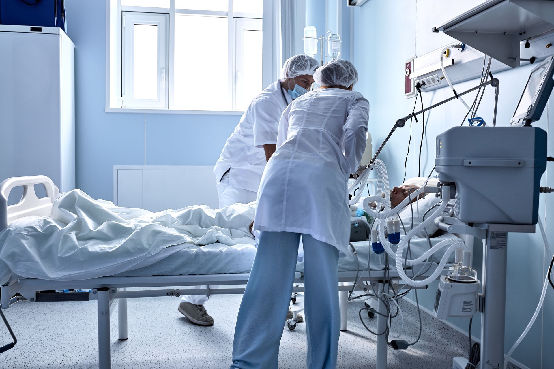 Deux professionnels de santé en blouse et charlotte s'occupent d'un patient dans une chambre de soins intensifs
