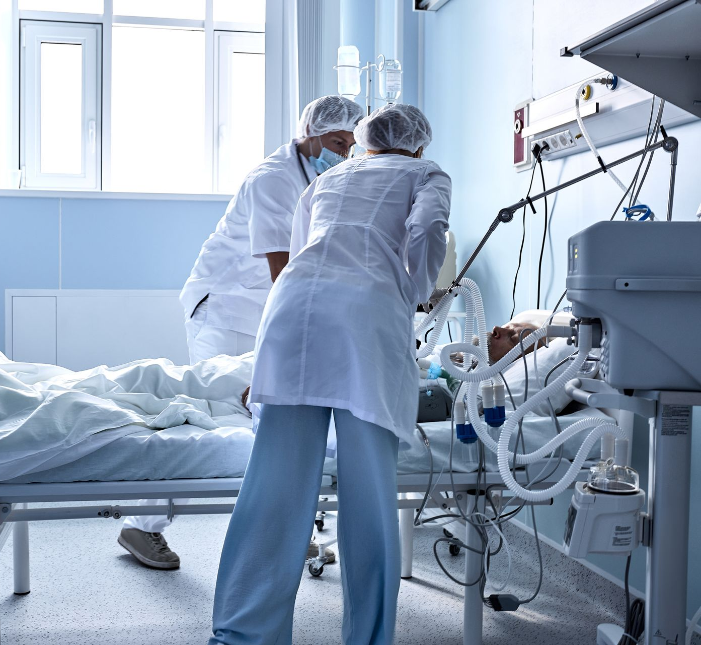 Deux soignants en blouse et charlotte s'occupent d'un patient en soins intensifs.
