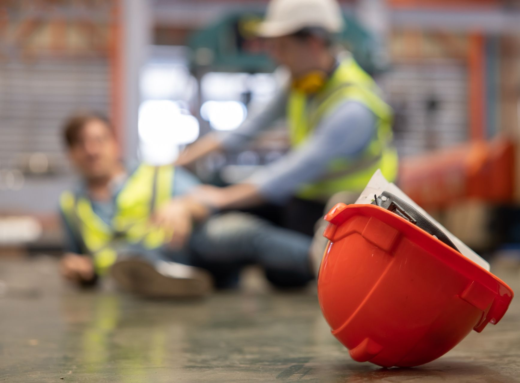 Au premier plan, un casque de chantier orange est posé sur un sol en béton, avec deux ouvriers du bâtiment derrière lui.