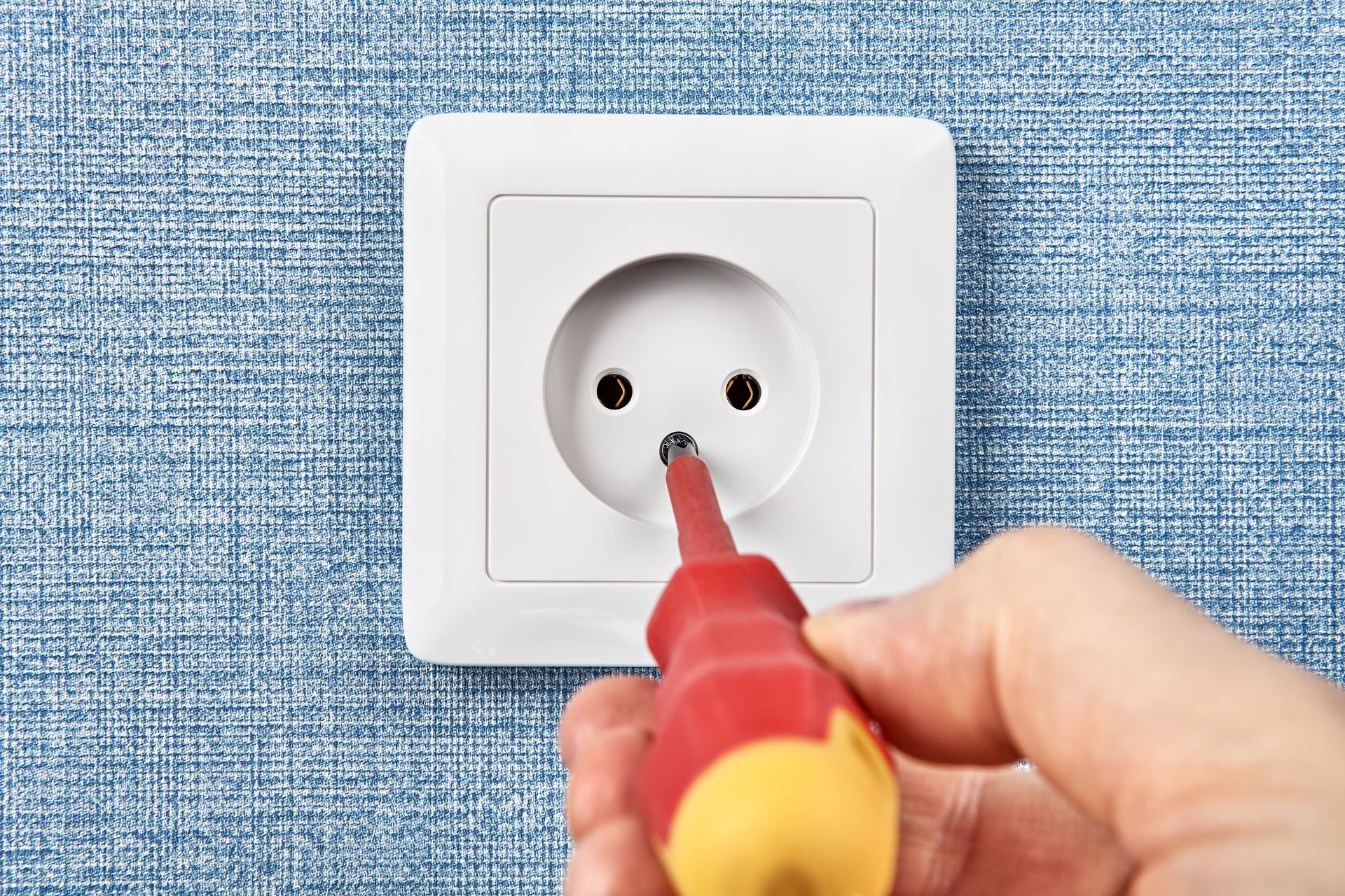 Main utilisant un tournevis sur une prise électrique blanche encastrée dans un mur texturé bleu.