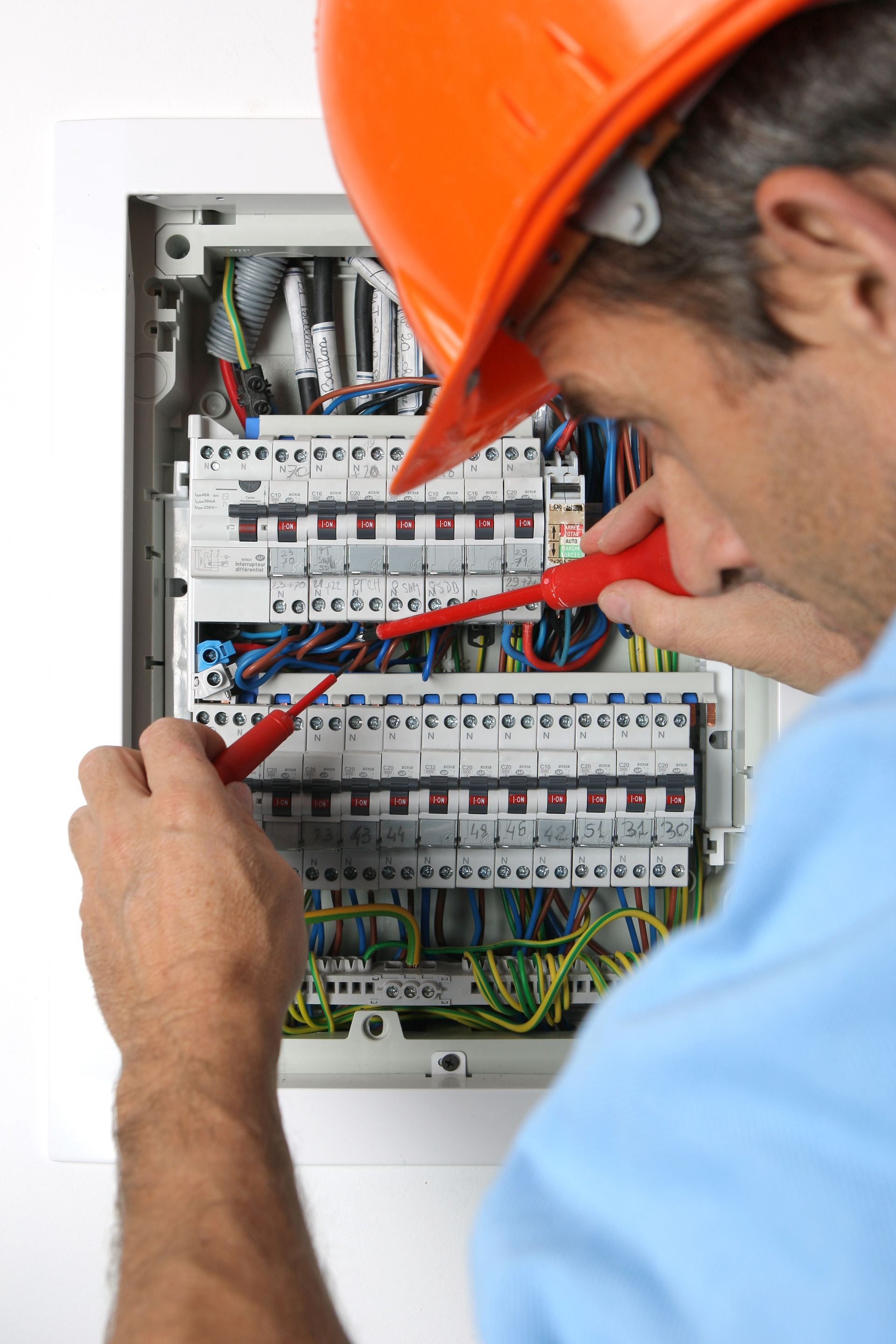 Un électricien portant un casque orange travaille sur un tableau électrique avec un tournevis.