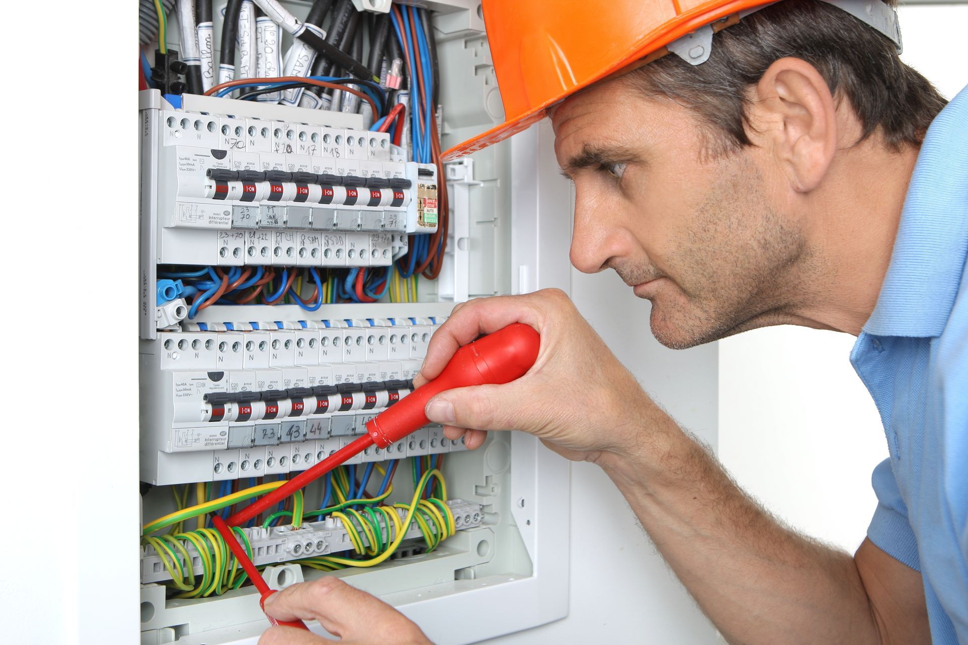 Un électricien portant un casque orange travaille sur un tableau électrique avec un tournevis.