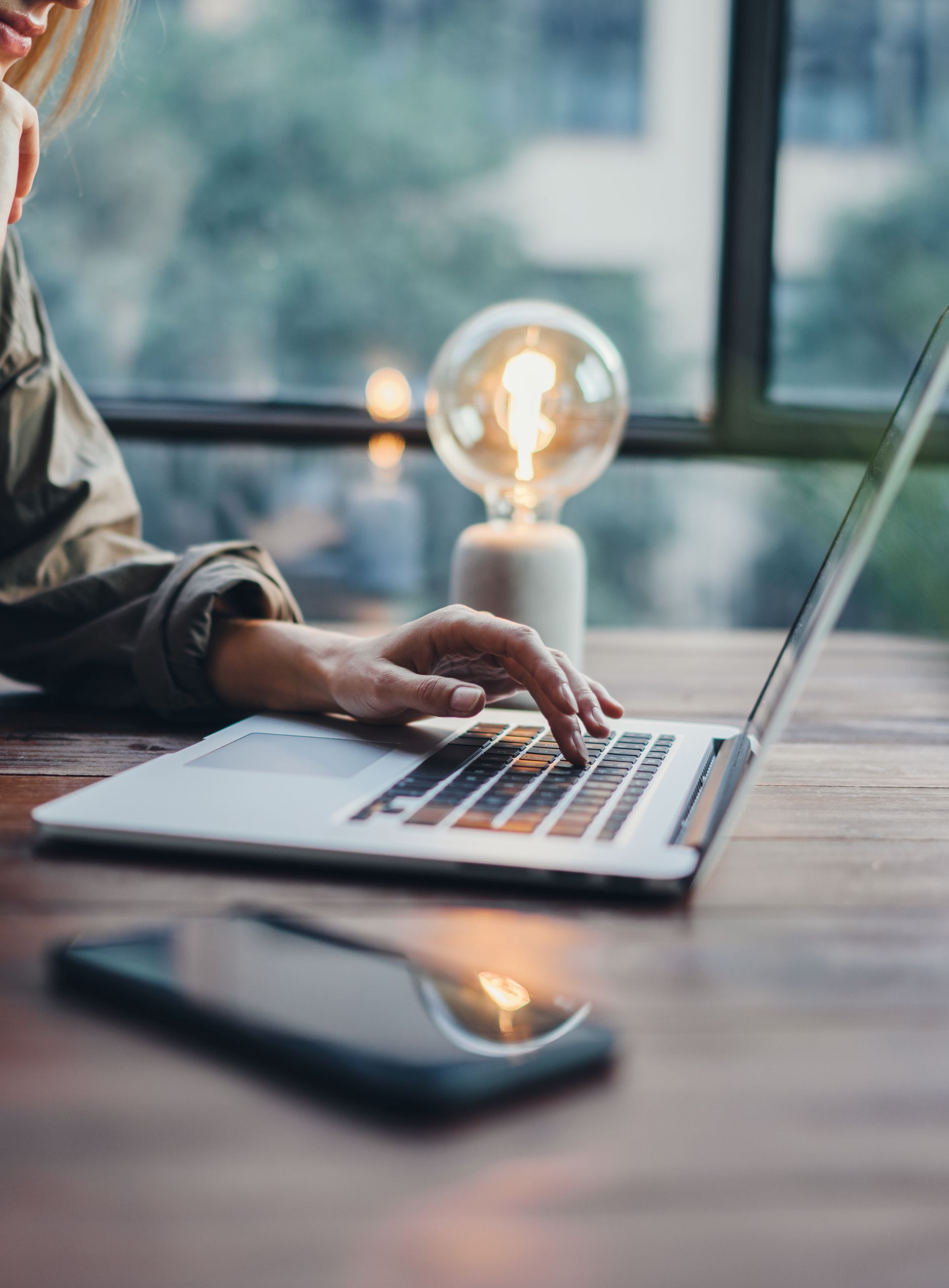 Femme tapant sur un ordinateur portable à une table en bois, smartphone au premier plan, ampoule allumée derrière.