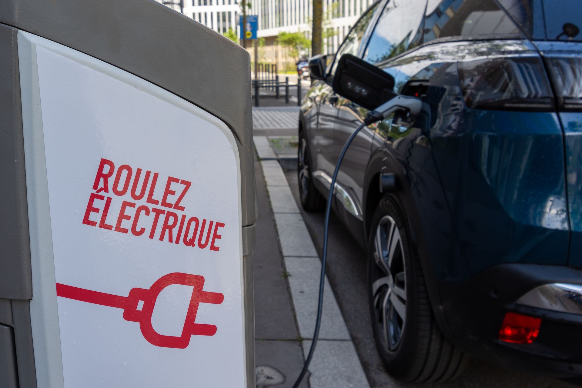 Voiture électrique en charge à une borne. Le panneau indique Roulez électrique avec un pictogramme de prise de recharge.