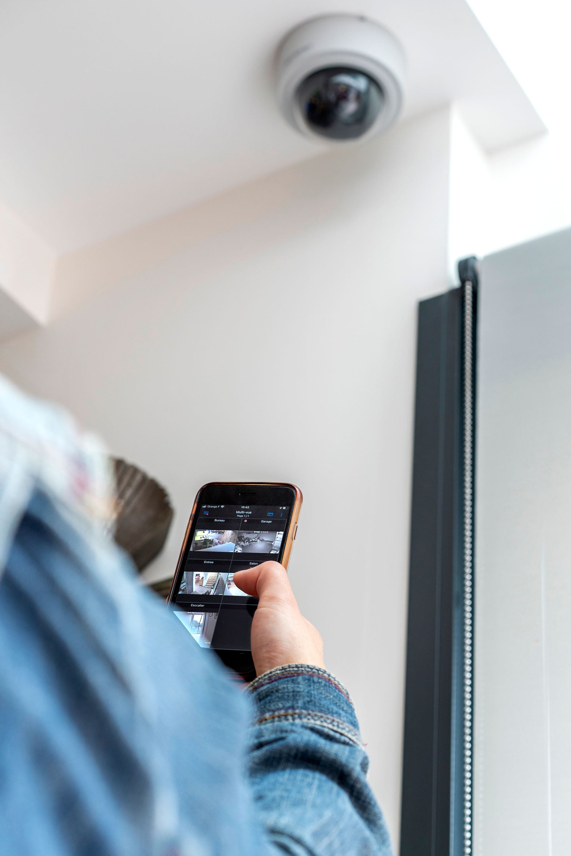 Une personne en veste en jean regarde les images d'une caméra de sécurité sur son téléphone, sous une caméra dôme fixée au plafond blanc.