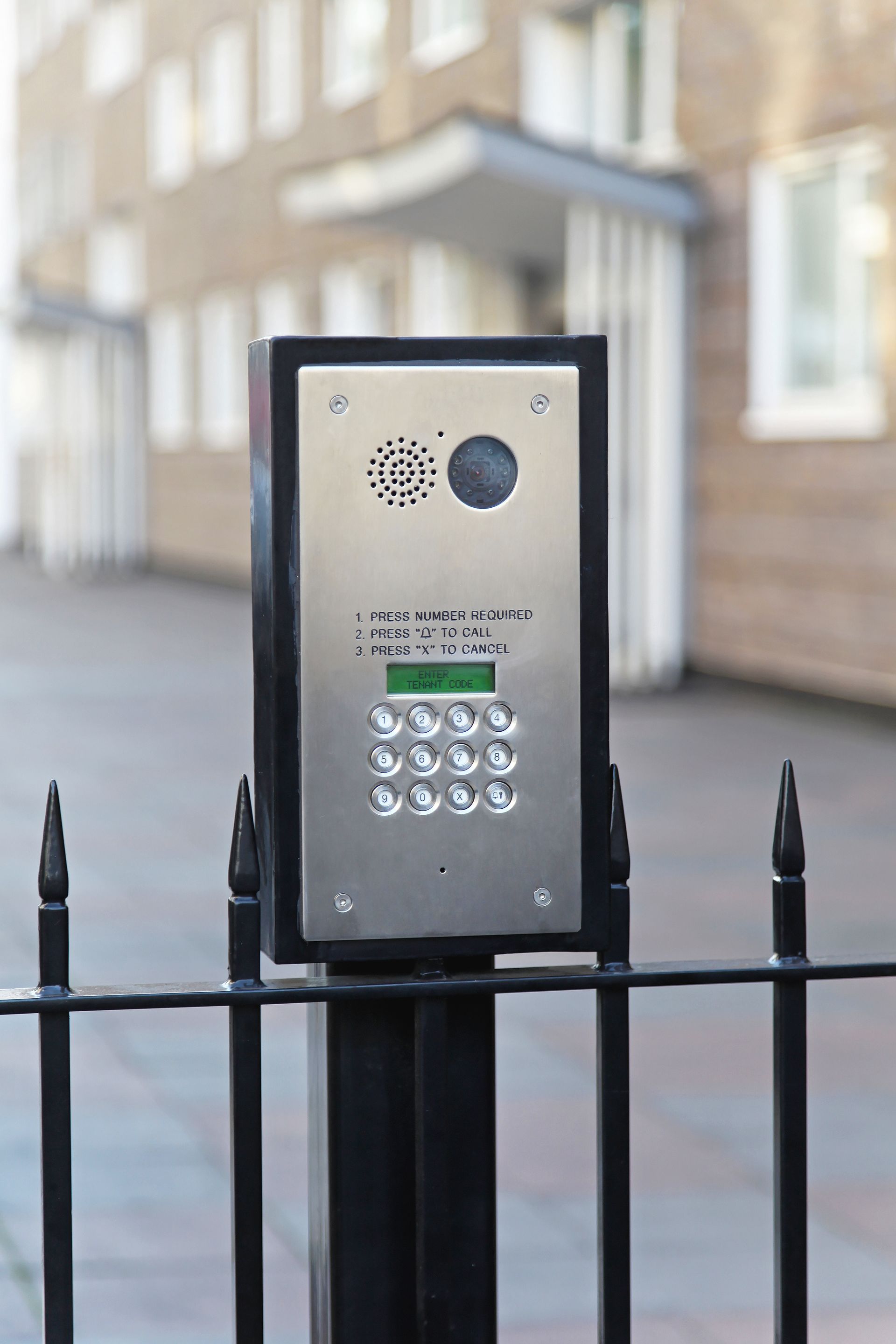 Un interphone argenté avec clavier, fixé sur un poteau noir derrière une clôture noire.