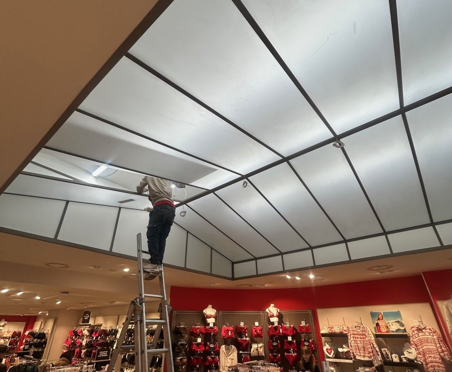 Dans un magasin, une personne perchée sur une échelle répare une partie d'un plafond blanc à bordures noires.