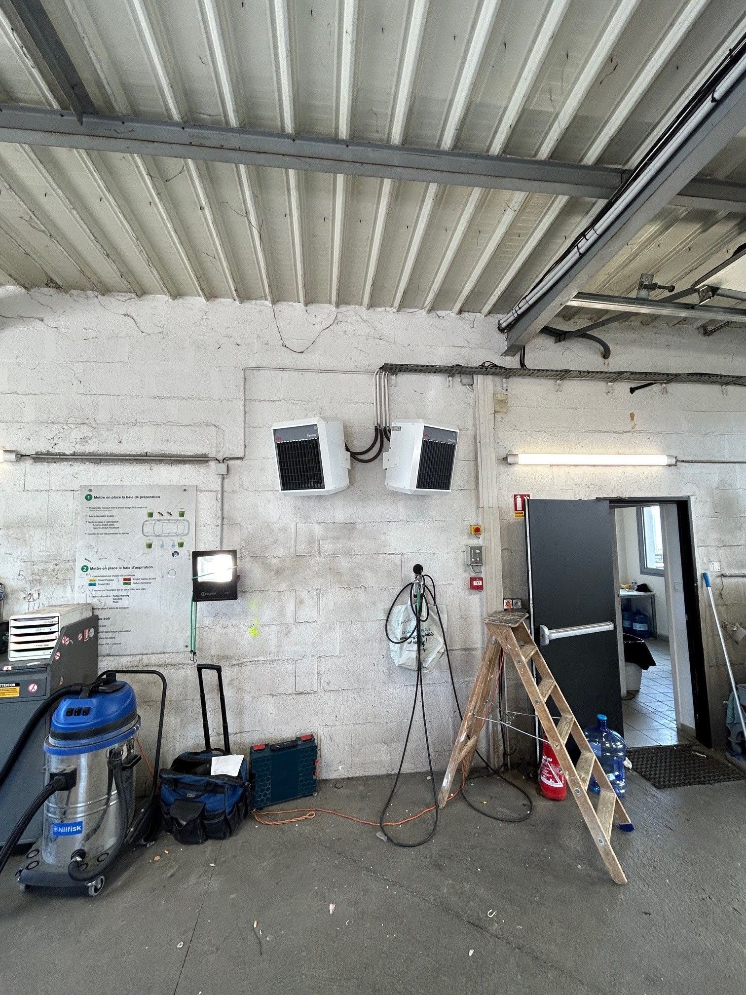 Deux purificateurs d'air fixés sur un mur de briques blanches dans un garage. Outils et équipements éparpillés sur le sol.