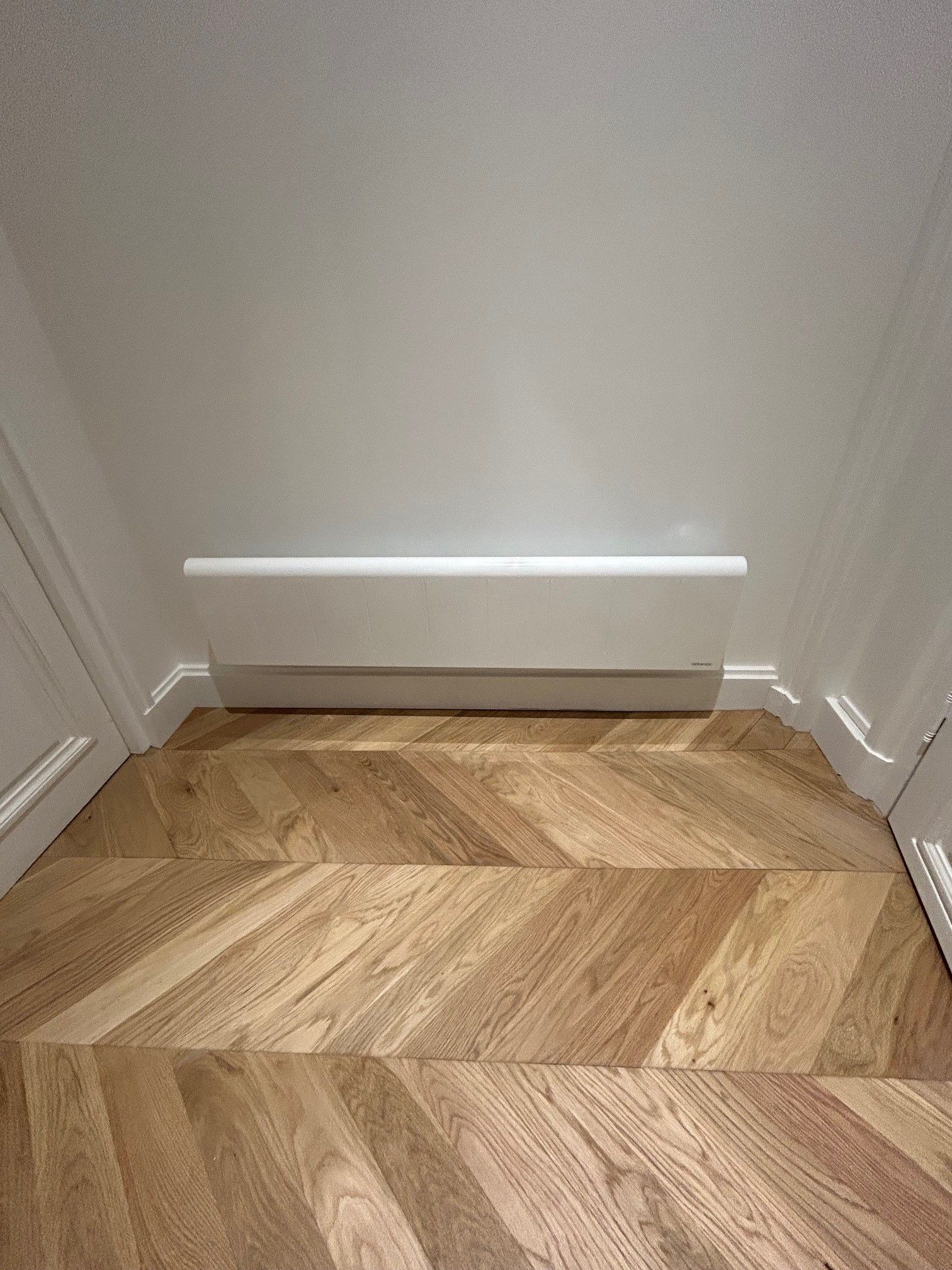 Sol en chevrons de bois, mur blanc et étagère blanche.