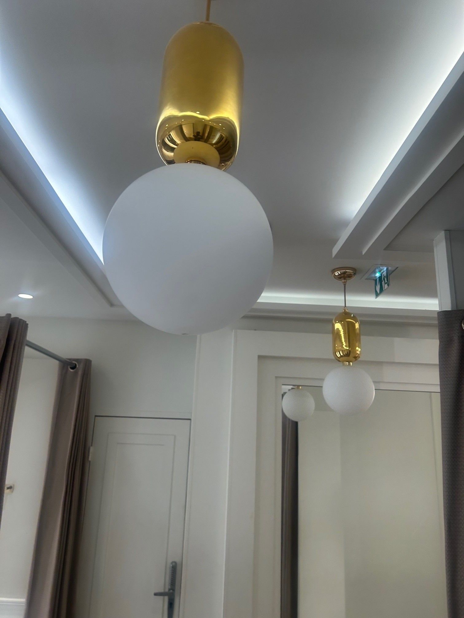 Des suspensions lumineuses dorées et blanches ornent un plafond blanc, au-dessus d'une pièce avec des portes et des rideaux.