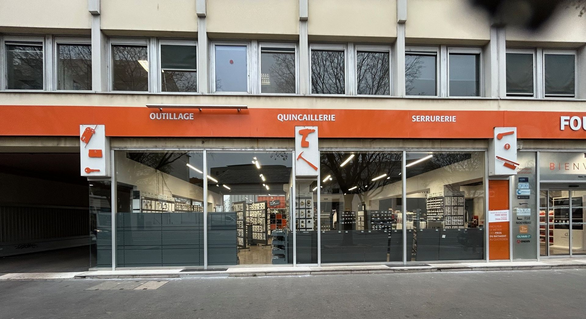 Magasin à enseigne orange, outils en vitrine, inscription OUTIL'SCIA. Extérieur du bâtiment, de jour.