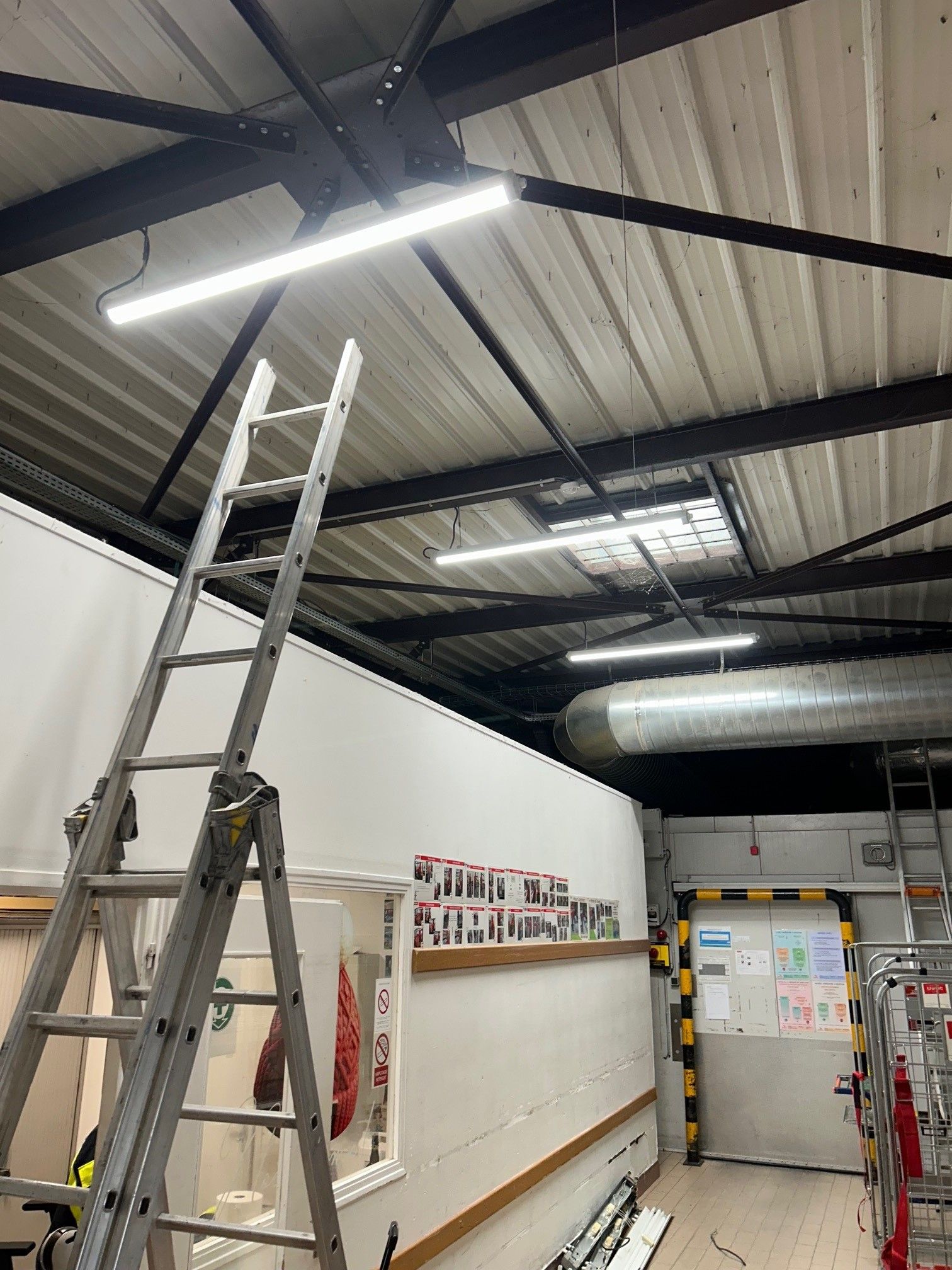 Échelle sous des néons dans un entrepôt. Murs blancs, plafond métallique.