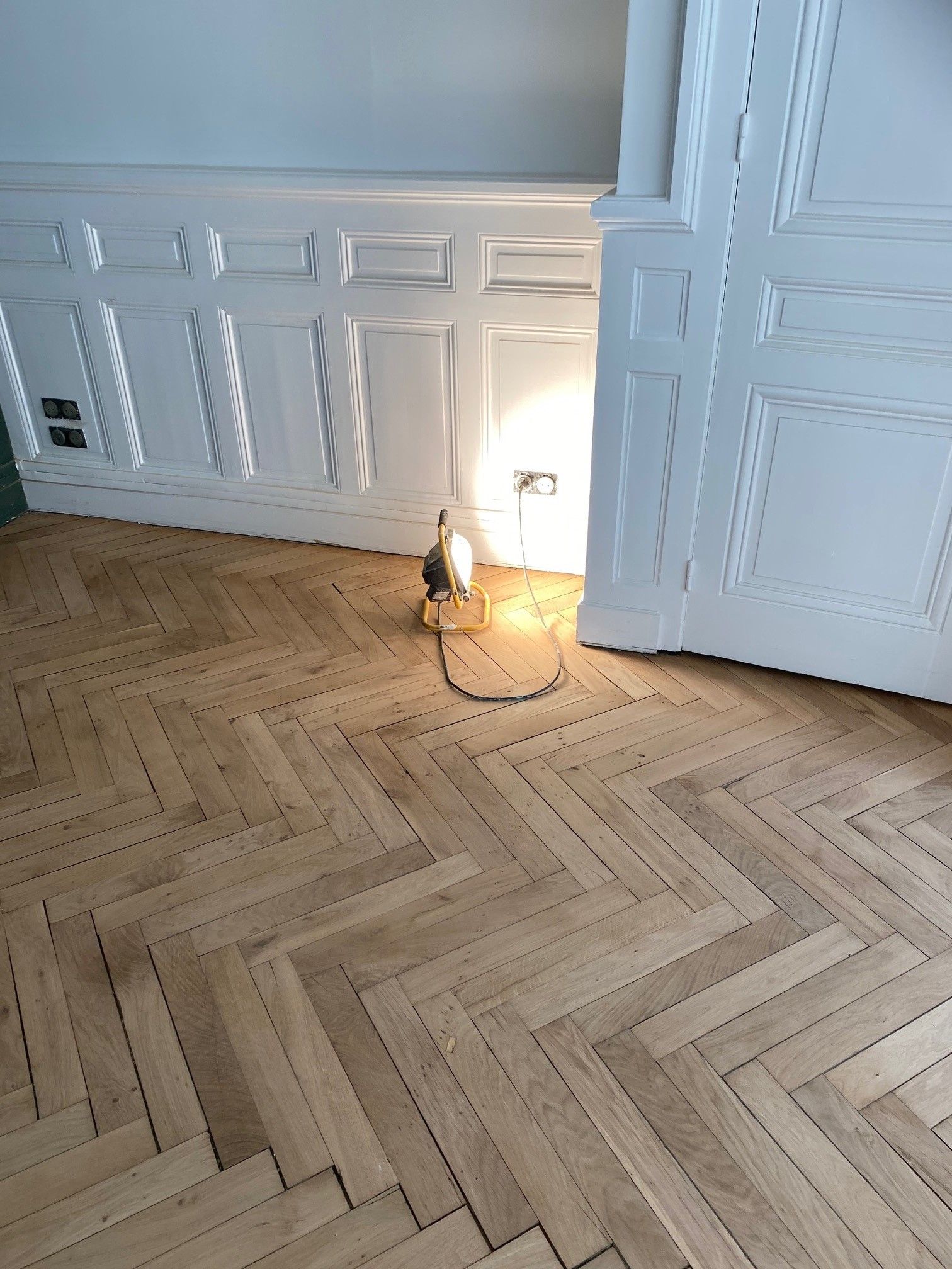 Parquet à chevrons, mur lambrissé blanc, petite lampe éclairant le sol.