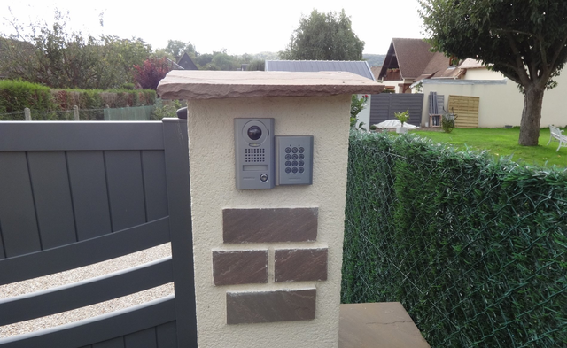 Pilier de portail gris avec interphone, clavier et brique décorative, surmonté d'une dalle de pierre.