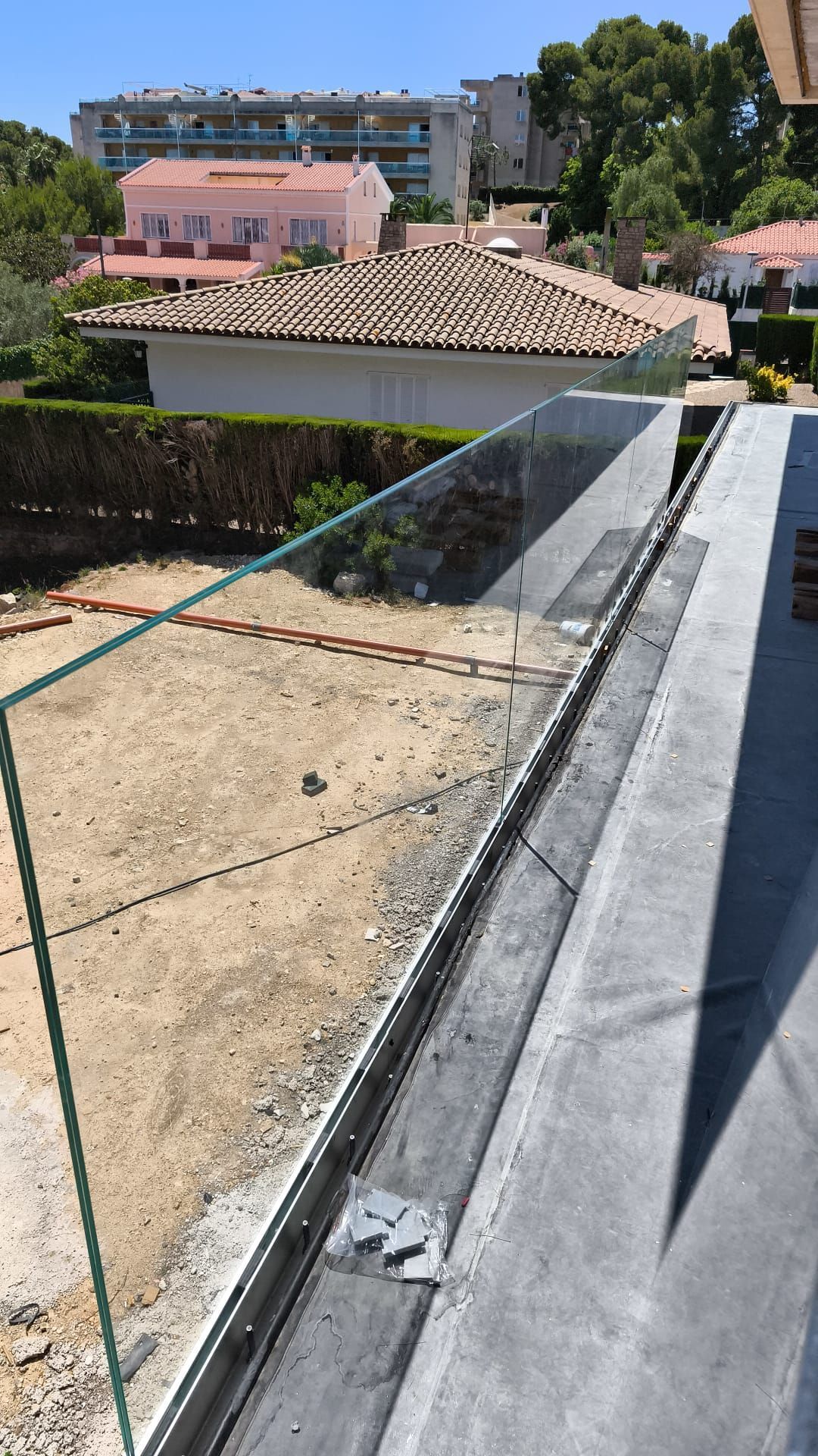 Barandilla de vidrio instalada sobre una superficie de hormigón, con materiales de construcción y edificios residenciales al fondo.