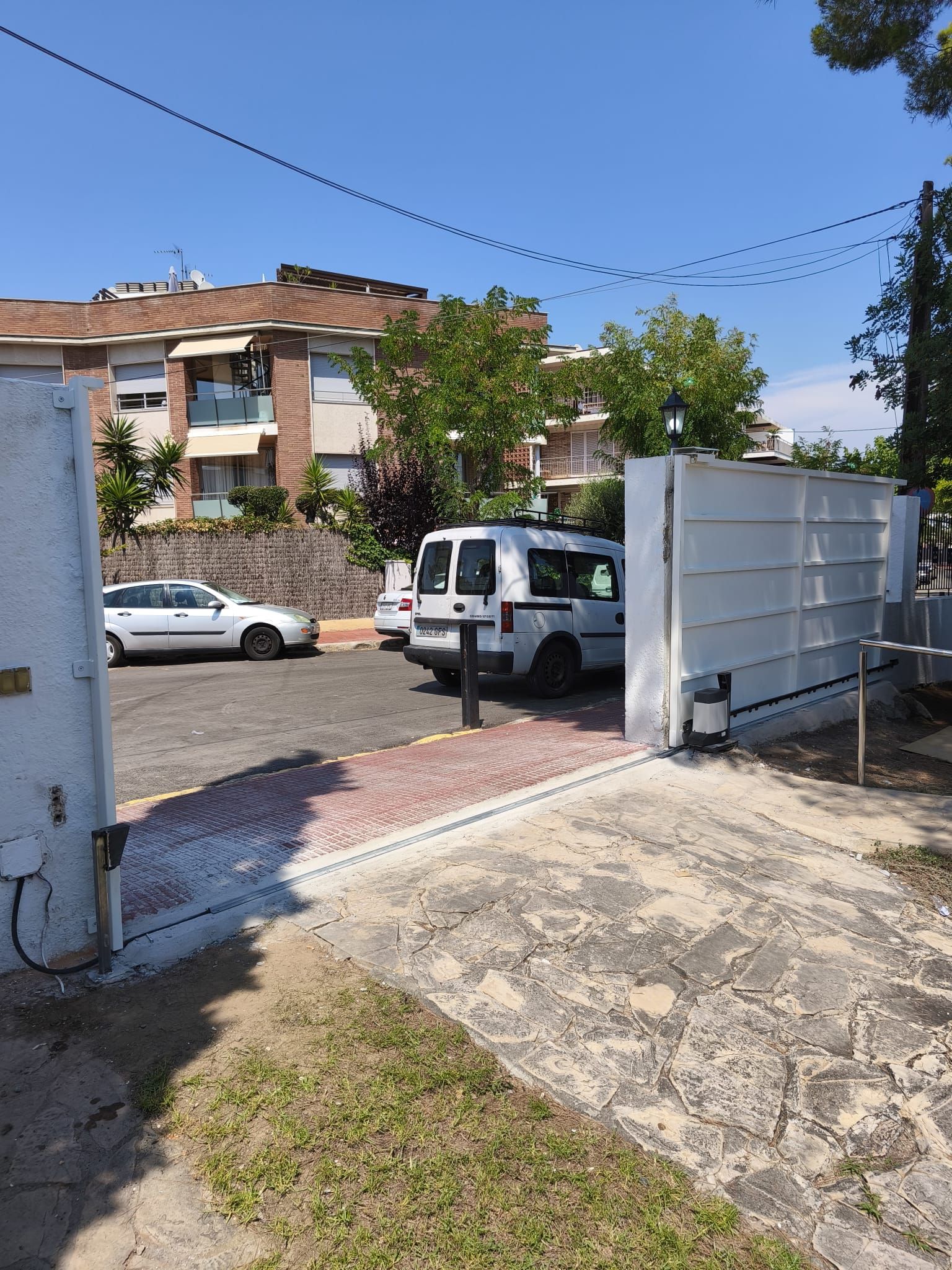 Puerta blanca abierta que conduce a un área de estacionamiento con una camioneta blanca, automóviles y un edificio en un día soleado.