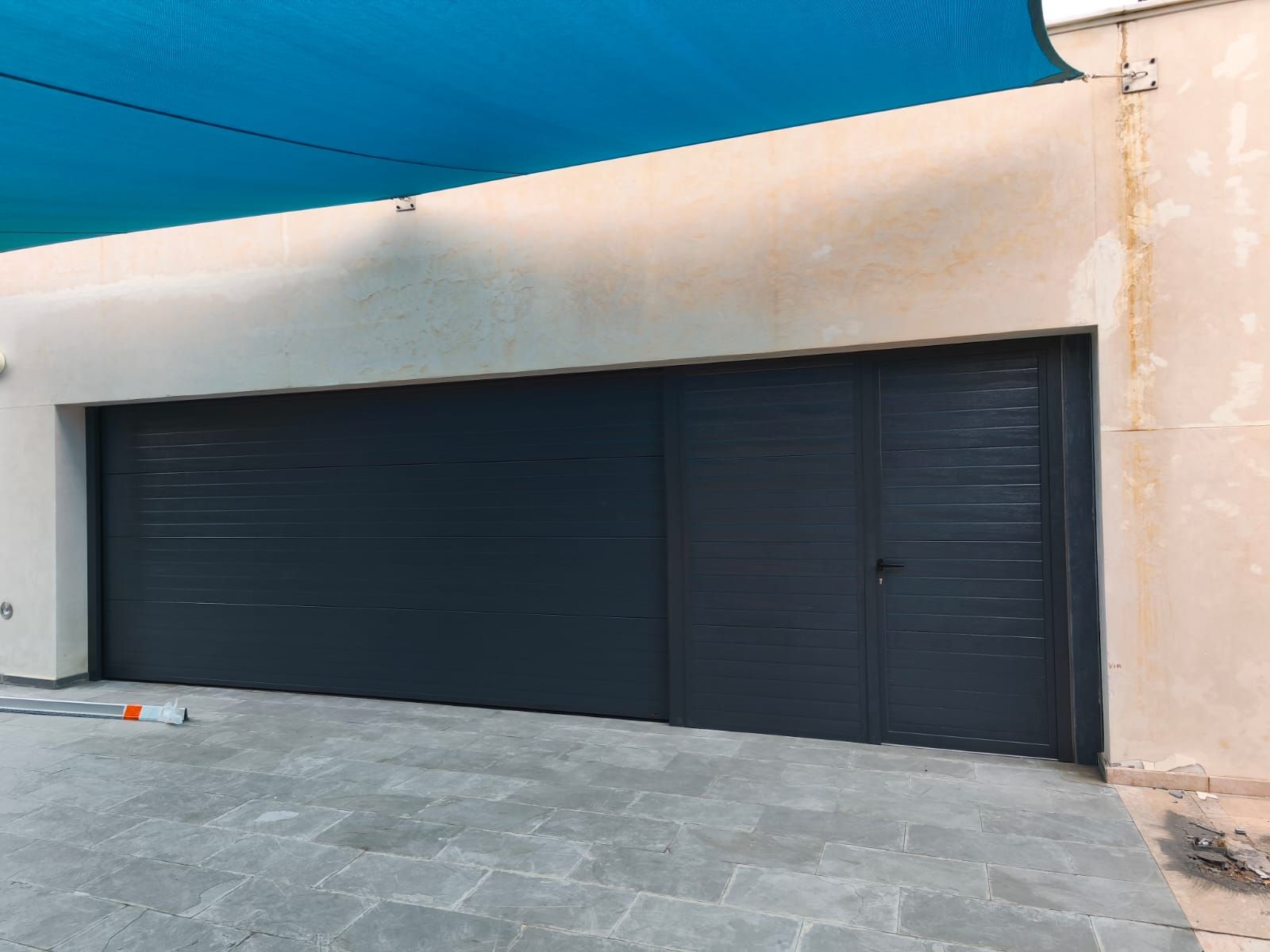 Puerta de garaje gris con puerta lateral, en contraste con un edificio de color claro y una pantalla azul.