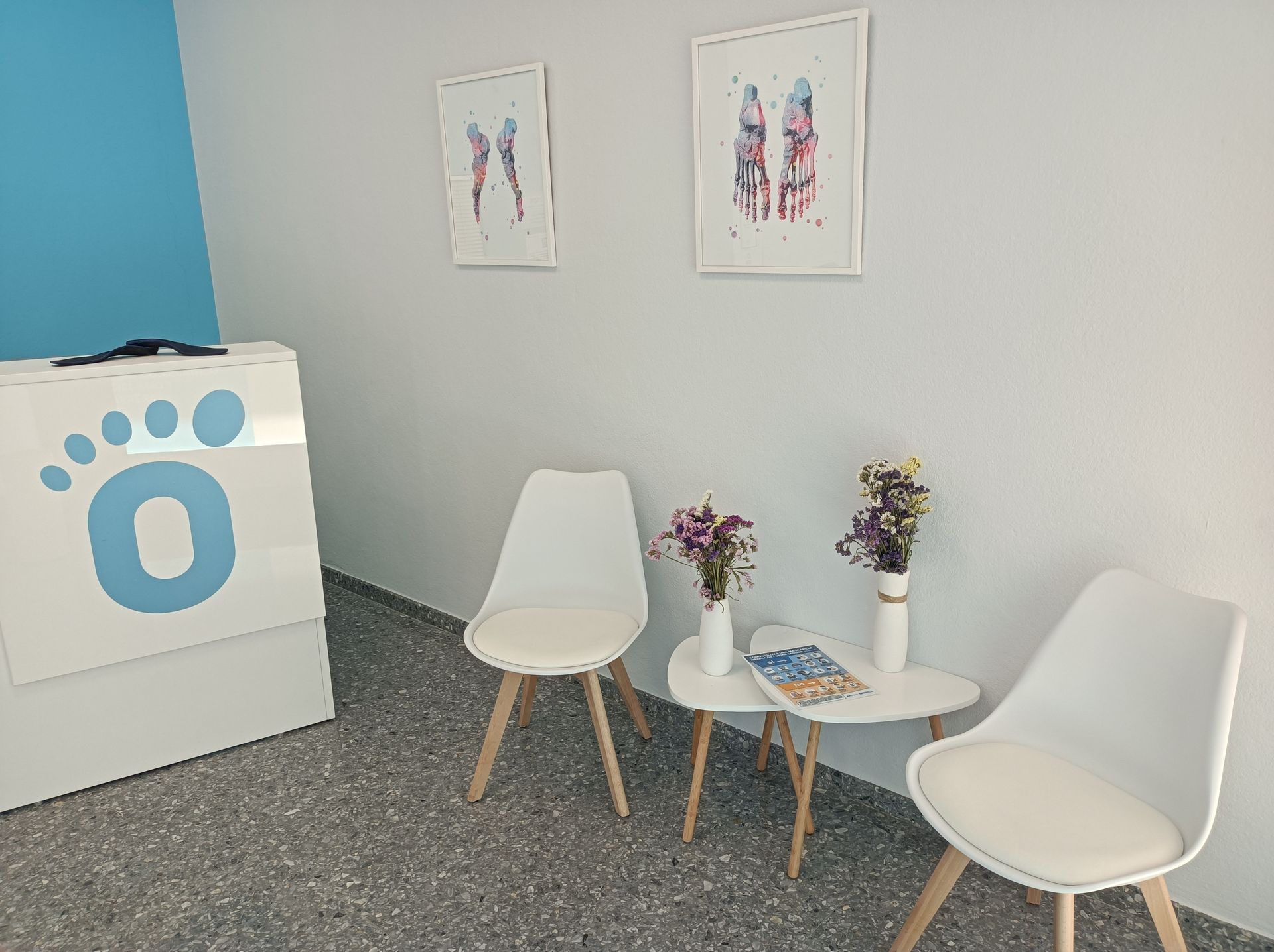 Sala de espera con sillas blancas, mesa pequeña, flores y logotipo en forma de pie; arte anatómico enmarcado.