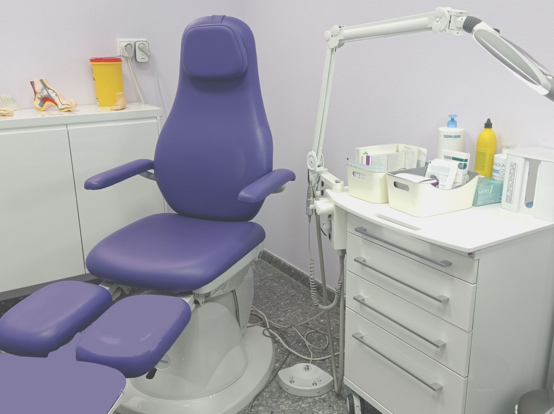 Silla médica de color morado en un consultorio médico bien iluminado, con un pequeño armario y una mesa de trabajo.