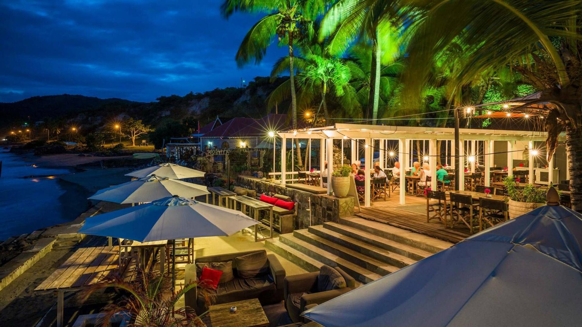 Een restaurant op het strand 's avonds met parasols en tafels.