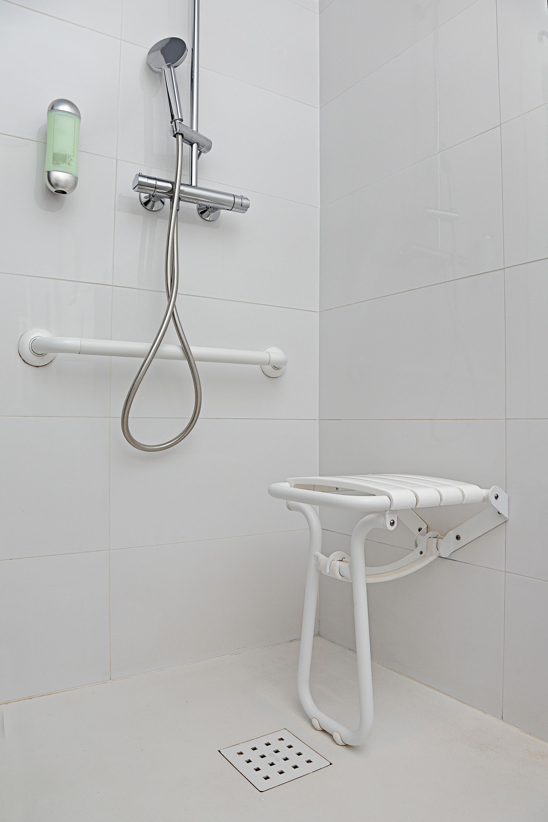 Douche accessible carrelée blanche avec siège rabattable, barre d'appui et pommeau de douche à main.