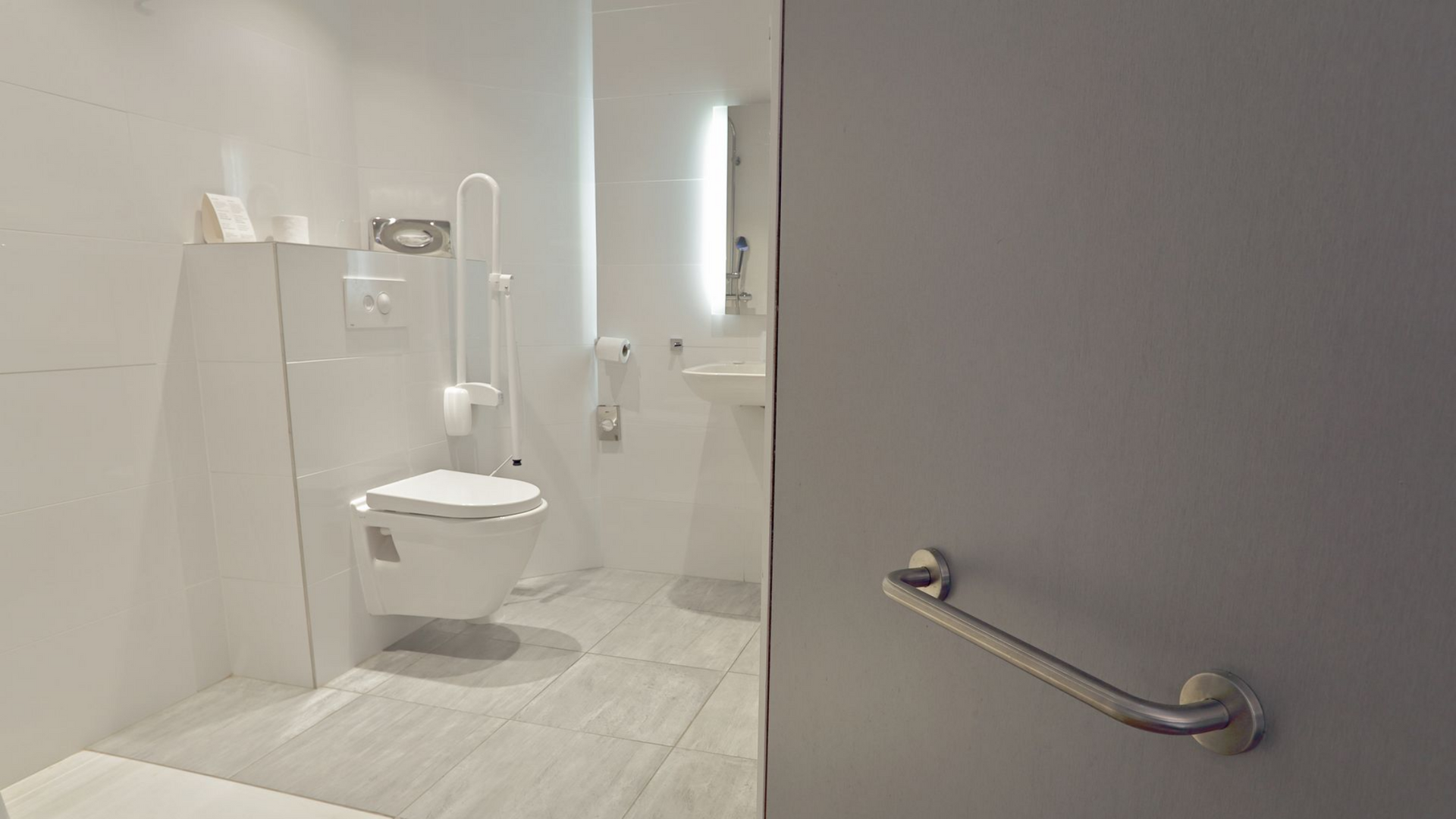 Salle de bains accessible avec murs blancs, toilettes, barres d'appui et porte grise partiellement visible.