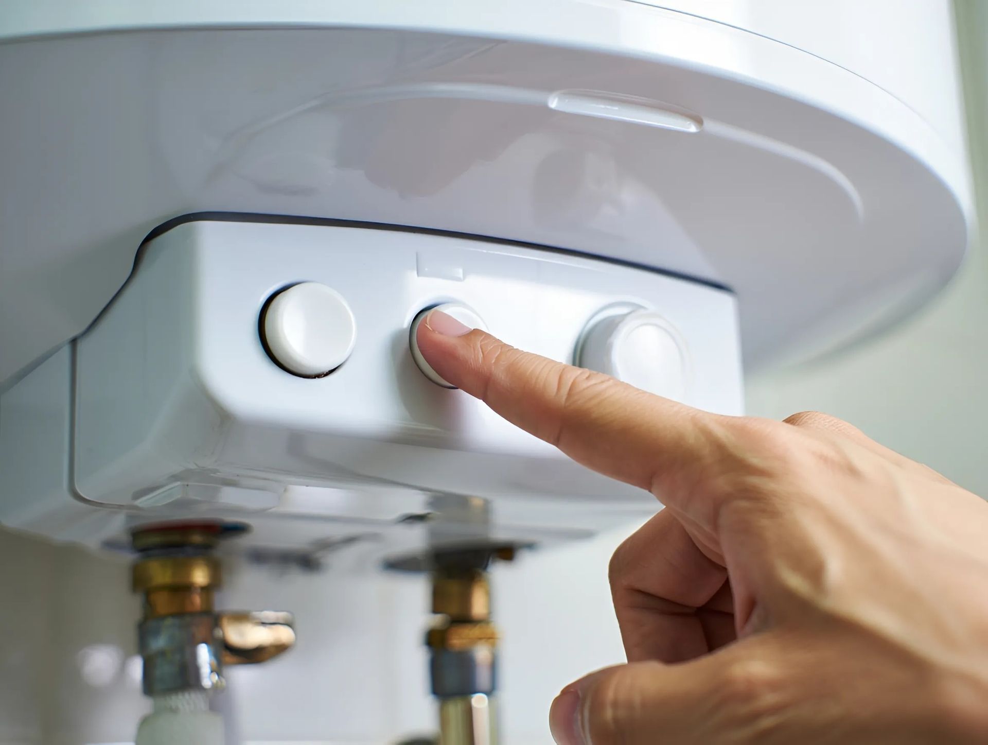 Main appuyant sur un bouton d'un chauffe-eau blanc, à proximité de tuyaux avec raccords en laiton.