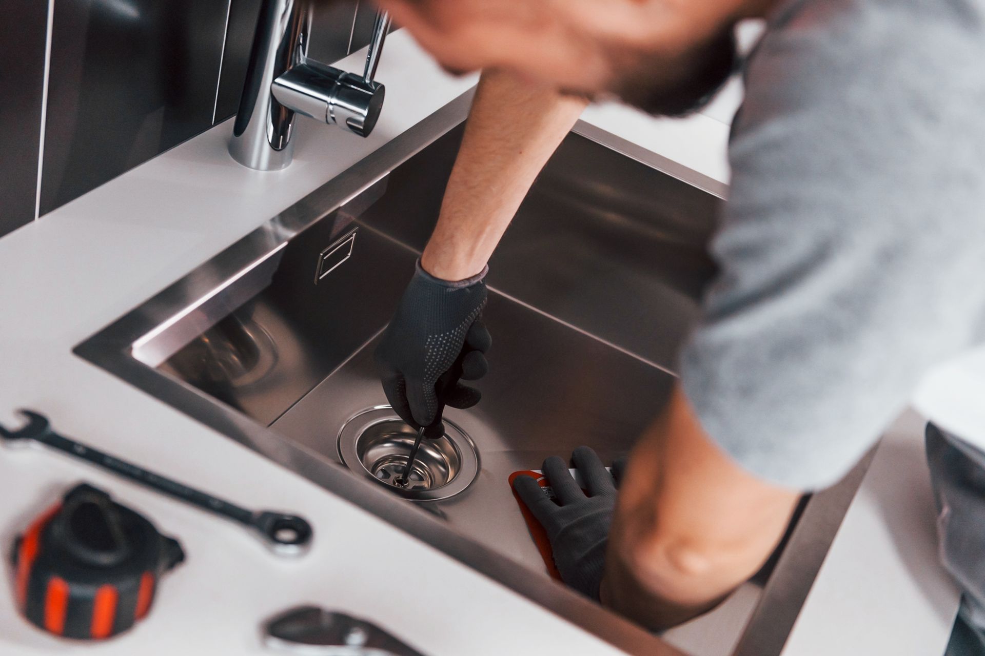 Plombier en gants noirs travaillant sur un évier de cuisine en acier inoxydable, avec des outils sur le comptoir.